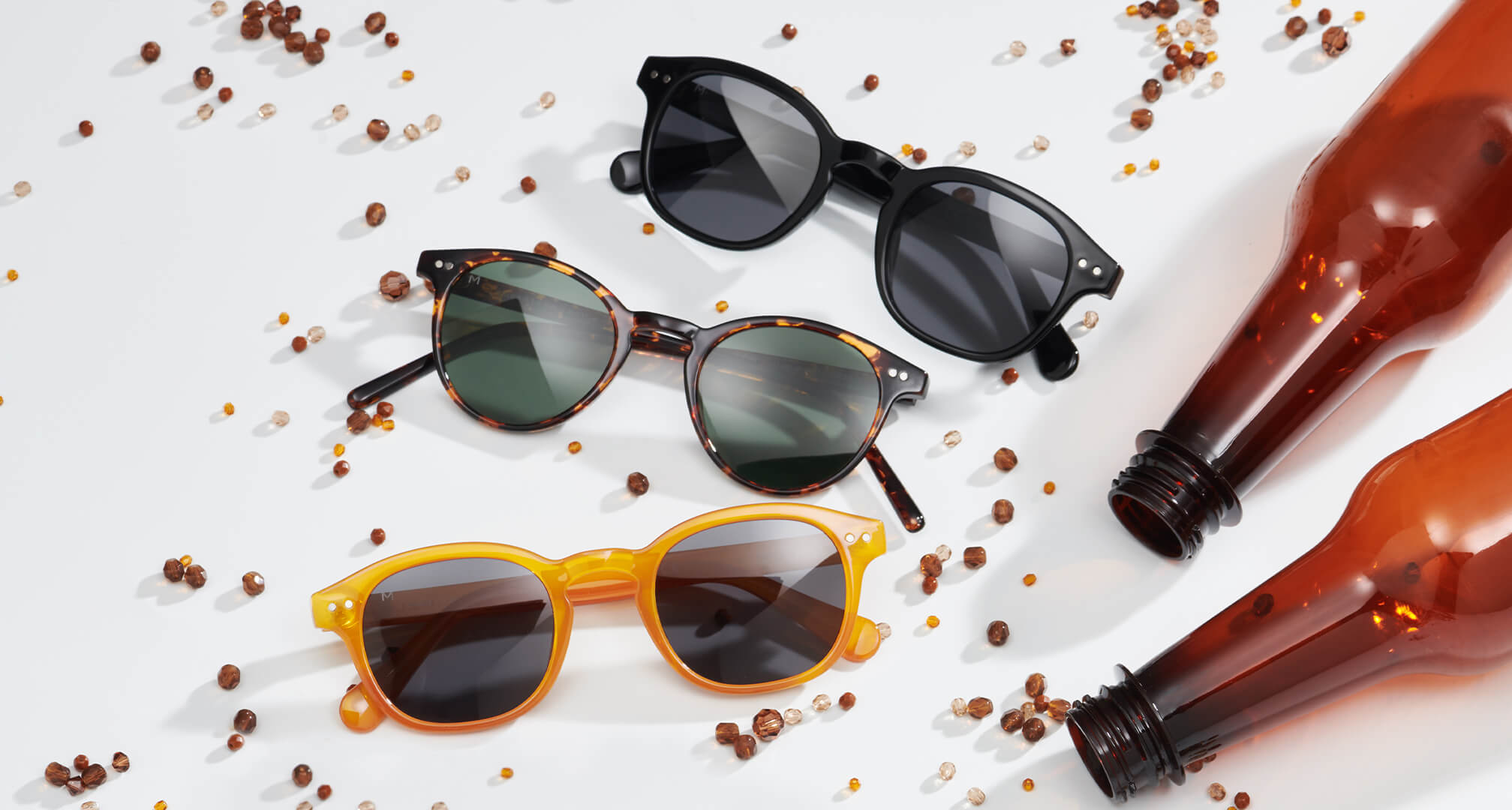 três pares de óculos de sol fabricados com materiais sustentáveis sobre uma mesa branca, com contas e garrafas de plástico à sua volta